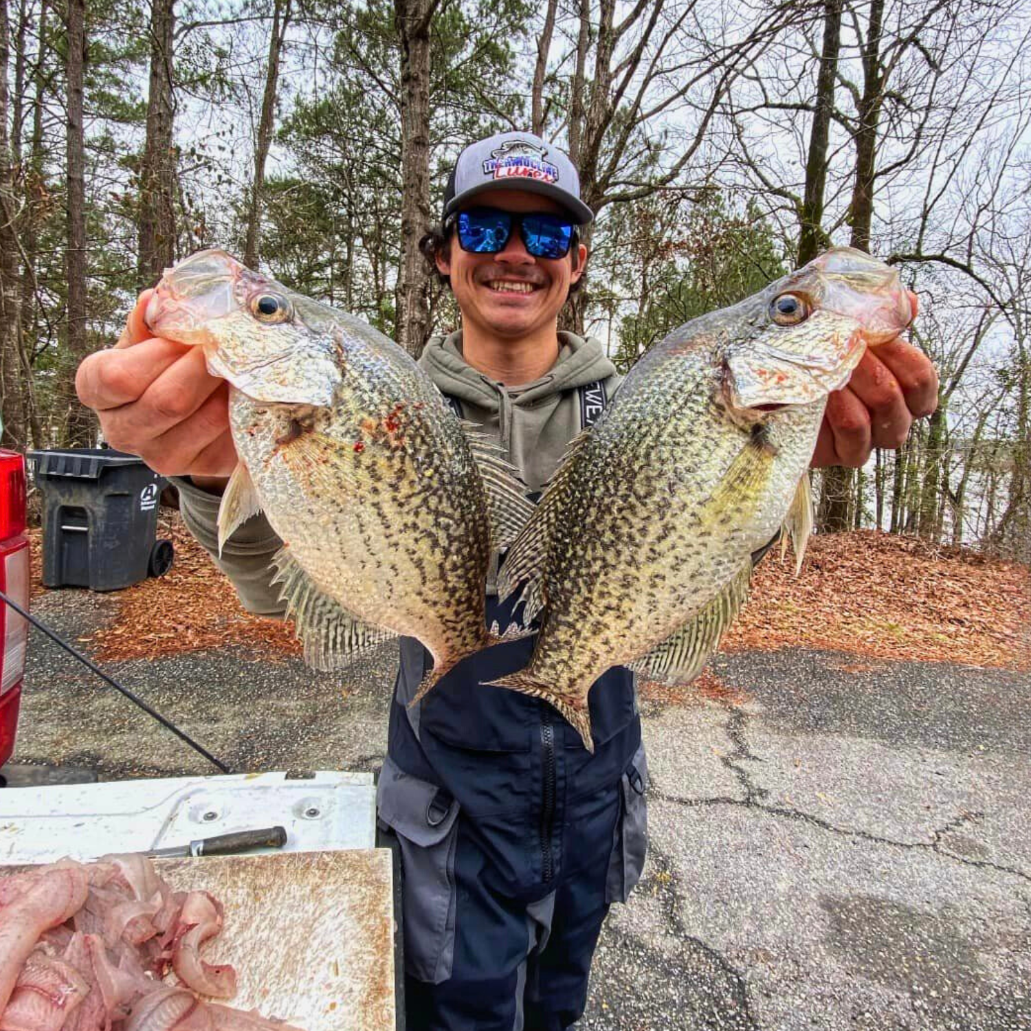 Blake holding two large crappie fish with a big smile, wearing a Thermocline Lures cap and standing outdoors in a wooded area.