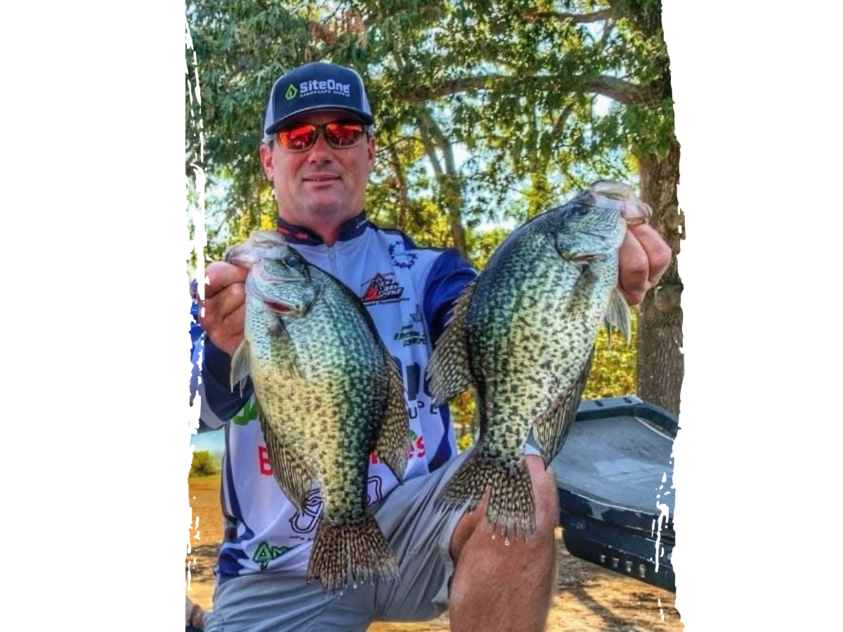 Clay Gann holding two crappie fish with a vibrant green and wooded background, dressed in a tournament fishing shirt and cap