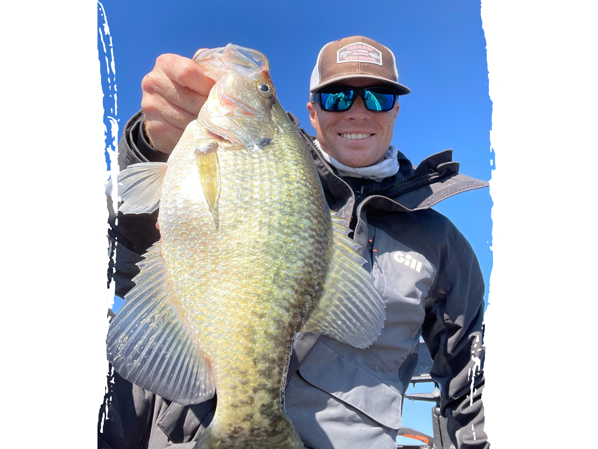 Dillon Gossett holding a golden crappie fish with a bright blue sky in the background, dressed in a fishing jacket and Thermocline trucker hat.
