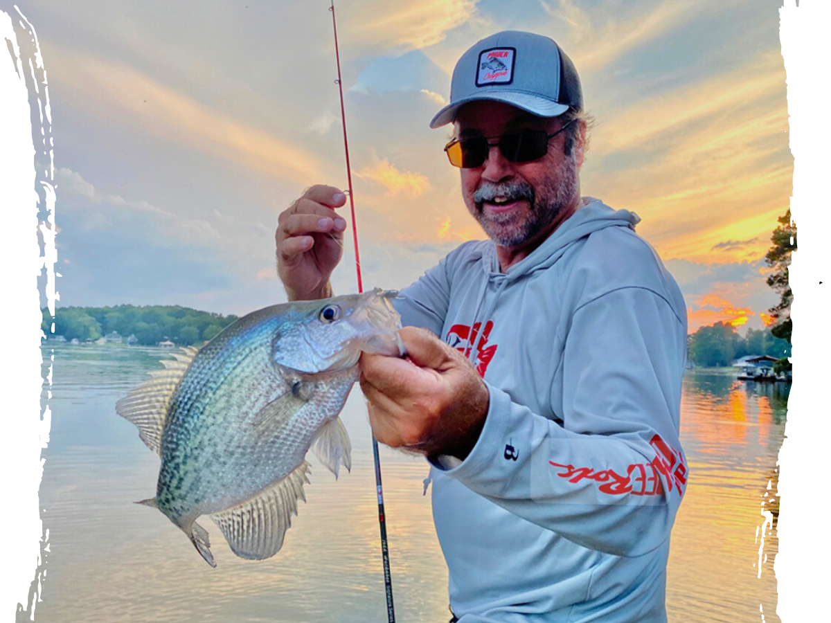 Joe Partenza holding a crappie fish near a serene lake backdrop, wearing sunglasses and a light gray fishing hoodie.